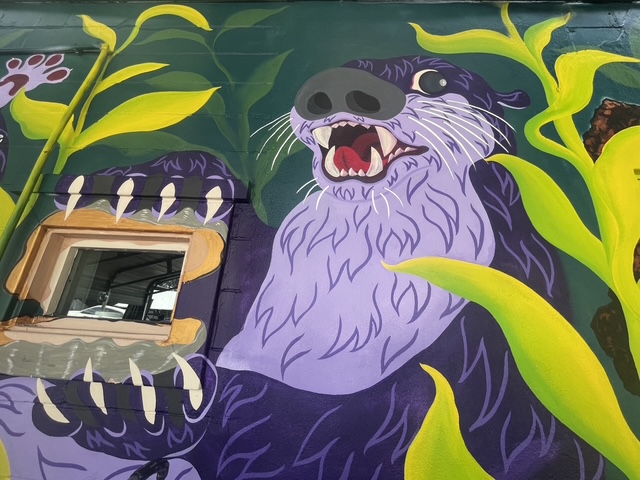 Colorful mural of a playful purple otter holding a clam among bright green leaves.