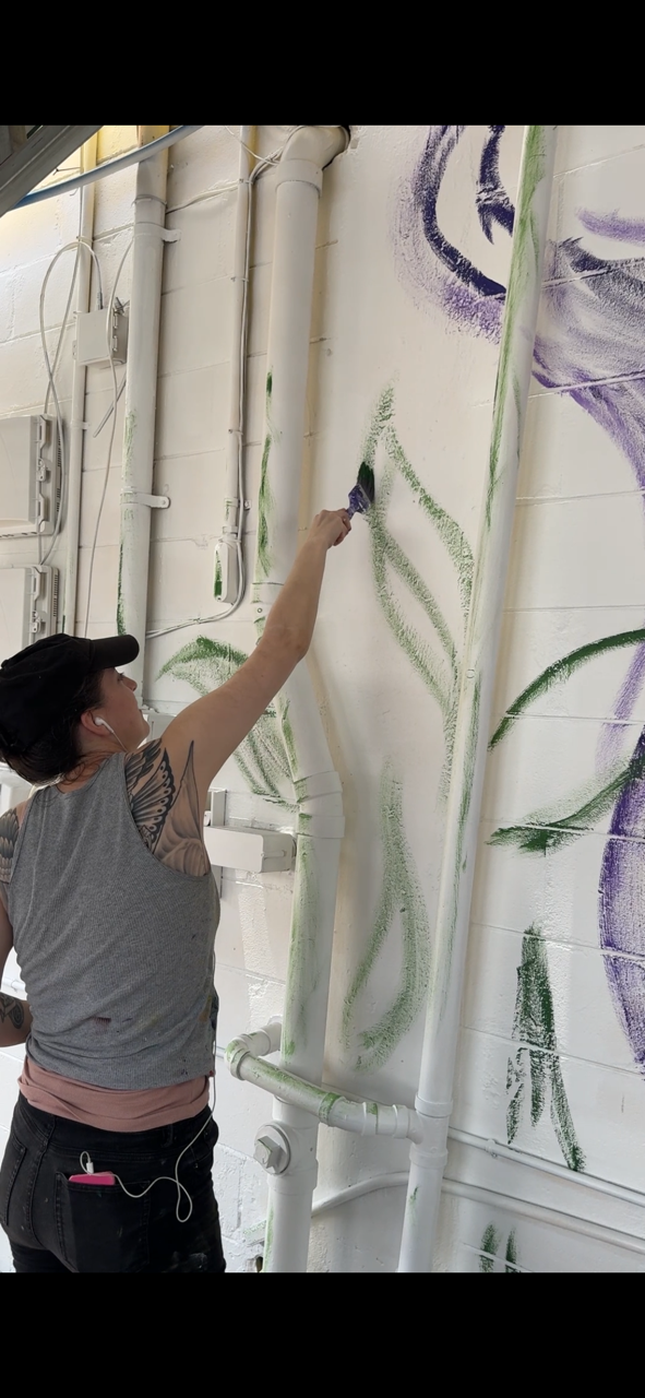 Minnie painting green leaf shapes on a white wall with purple mural details nearby.