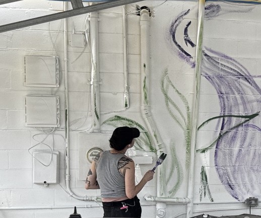Minnie standing in front of an in-progress wall mural with green leaves and purple forms.